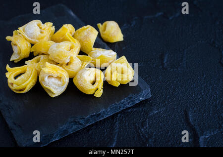 Frisch hausgemachten raw flouring Tortellini bereit, auf dunklen Stein Platte auf schwarzen Hintergrund zu Koch zubereitet. Traditionelle italienische Küche. Kopieren spa Stockfoto