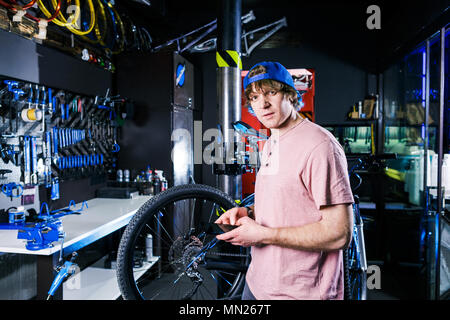 Eine junge, elegante rothaarigen Mann Small Business Owner verkauf und reparatur ein Fahrrad trägt eine blaue Kappe und ein rosa Trikot ist mit einem Handy whil Stockfoto