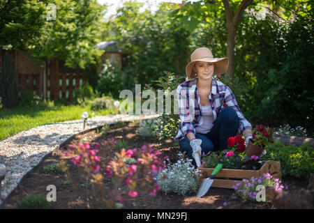 Schöne weibliche Gärtner an Kamera suchen und lächelnd Holzkiste voller Blumen in Ihrem Garten gepflanzt werden. Gartenarbeit Konzept. Stockfoto
