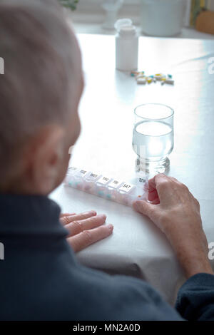 Alte Frau an einem Tisch zu Hause sitzen mit einem Glas Wasser und viele Kapseln in verschiedenen Farben in einer Vorsortierten wöchentliche Tablettenspender. Stockfoto