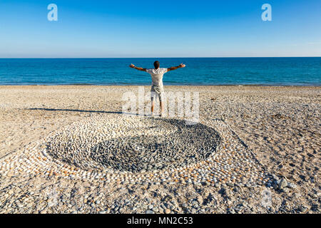 Erde Kunst am Strand von Palavas-les-Flots (Südfrankreich): Yin und Yang Symbol mit Kieselsteinen am Strand und Mann gesehen von hinten, Arme erstellt Ausdrucke Stockfoto