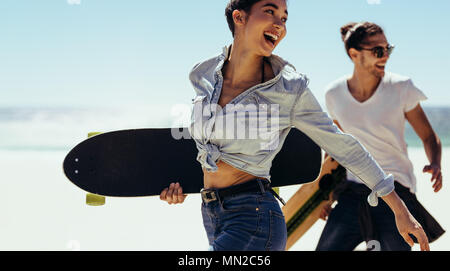 Lächelnde Frau mit ihrem Freund am Strand holding Skateboards. Die jungen Skater zu Fuß am Strand. Stockfoto