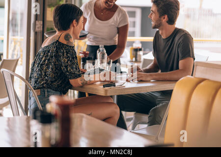 Junges Paar im Cafe sitzen, um eine weibliche Kellner. Mann platzieren Um eine Kellnerin im Restaurant sitzend mit seiner Freundin. Stockfoto