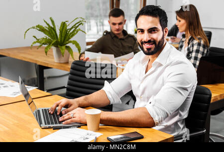 Attraktive bärtiger Mann im weißen Hemd arbeiten an Silber Laptop, zwei andere Mitarbeiter, die die Aufgabe hinter dem Mann Stockfoto