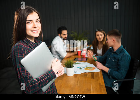Hübsche dunkelhaarige Frau mit Laptop im Büro, drei smart Manager diskutieren mögliche Lösungen am Schreibtisch im Hintergrund, Portraitfotos Stockfoto