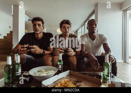 Freunde Spaß mit Getränken und Snacks beim Spielen von Spiel zu Hause. Zwei Männer spielen video game Holding Joysticks, während ein anderer Mann fei Uhren Stockfoto