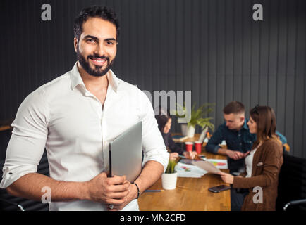 Gerne asiatischer Mann mit schwarzen Bart holding Silber Notebook, stehen vor dem Kollegen arbeiten an der Aufgabe, die Vorbereitung der nächsten Phase des Projekts im Büro Schreibtisch mit Diagrammen, Kaffeetassen und Gadgets Stockfoto