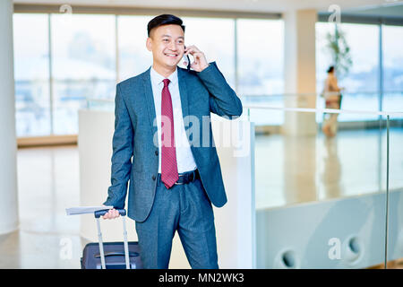 Portrait von modernen asiatischen Geschäftsmann sprechen durch Telefon und lächelt glücklich während der Wartezeit in der Airport Holding Koffer Stockfoto
