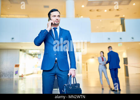 Portrait von Stattlichen elegante businesssman sprechen Telefonisch holding Gepäck in modernen Flughafen, Kopie Raum Stockfoto