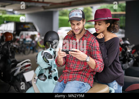 Happy interracial Paare auf dem Parkplatz im Freien. Weißer Kerl in einem roten Hemd sitzt auf einem Motorrad und schaut auf sein Smartphone, schwarze Mädchen in einem crim Stockfoto