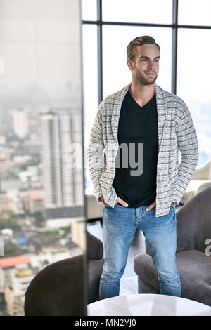 Hübscher Kerl steht in seiner Wohnung auf die großen Fenster im Hintergrund. Er trägt einen hellgrauen Cardigan, Schwarz T-Shirt und eine blaue Jeans. Mann hält seine Hände in Stockfoto
