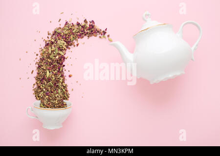 Trockene Kräuter Tee einzuschenken aus weißem Porzellan Teekanne in den Becher auf rosa Oberfläche. Flach. Tea time Konzept. Stockfoto