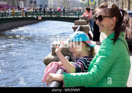 Junge Mutter mit der Tochter lächeln und willkommene Touristen im sonnigen Sommer und windigen Tag. Auf einem unscharfen Hintergrund Griboyedov Kanal in St. Petersburg Stockfoto