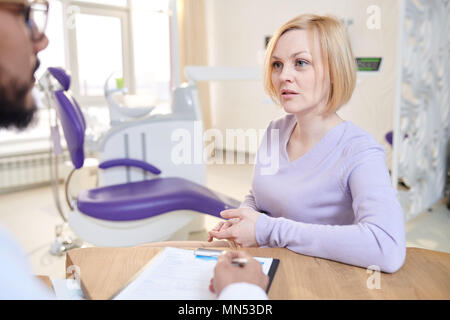 Portrait von besorgt Frau sprechen, sitzen am Schreibtisch in der modernen zahnmedizinischen Klinik und beschreibt Symptome zu Arzt Stockfoto