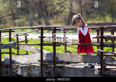 Die hübsche kleine Blonde langhaarige Mädchen in schönes rotes Kleid steht allein auf alten Zement Brücke gelehnt auf hölzernen Geländer versessen auf warmen sonnigen Stockfoto