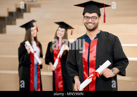 Glücklich lächelnde junge halten Universität Diplom. Absolvent in der Nähe von jungen groupmates und Suchen zufrieden. Die Studierenden tragen schwarz und rot gefärbte Graduierung Kleider. Ende der Universität. Stockfoto