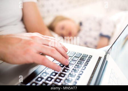 Nicht erkennbare Vater mit Laptop und einem schlafenden Kind Junge im Bett zu Hause. Vaterschaftsurlaub. Stockfoto