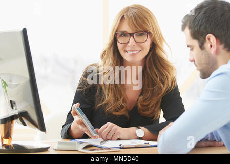 Im mittleren Alter Berater geschäftsfrau am Schreibtisch sitzen und mit Handy während Beratung mit jungen männlichen business Assistent im Büro. Stockfoto