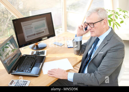 Hohe Aufnahme von Senior Sales Mann am Schreibtisch vor dem Laptop sitzt und spricht mit jemand in sein Mobiltelefon. Stockfoto