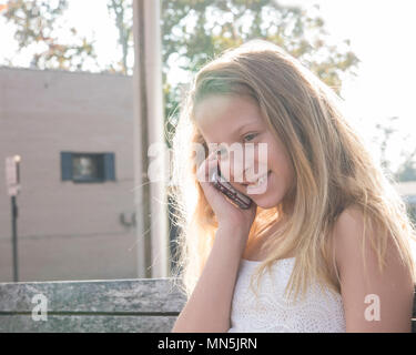 10 Jahre alte Mädchen sitzen auf einer Bank im Gespräch über Ihr Handy, ein Lächeln auf den Lippen. Stockfoto