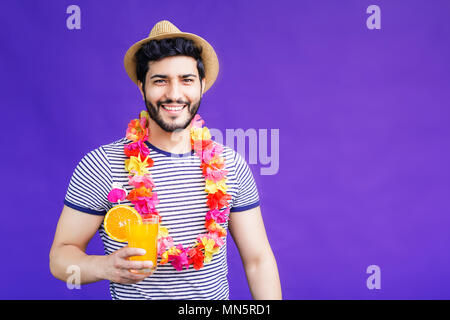 Gut aussehender bärtiger Mann in Lei auf Hals und Hut bekleidet und hält Orange Drink auf lila Hintergrund, Ferienhäuser Konzept, isolierte Studio shot Stockfoto