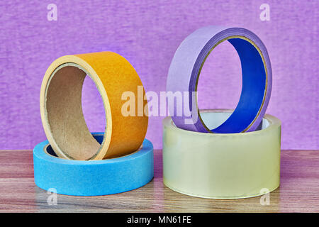 Vier Rollen Klebeband in verschiedenen Farben und für verschiedene Zwecke liegen auf einer Tabelle auf einem lila Hintergrund. Stockfoto