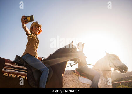 Paar auf Pferden reiten im Freien unter der Sonne im Gegenlicht für Freizeit Aktivitäten selfie mit modernen Mobilfunktechnologie. Alternative li Stockfoto