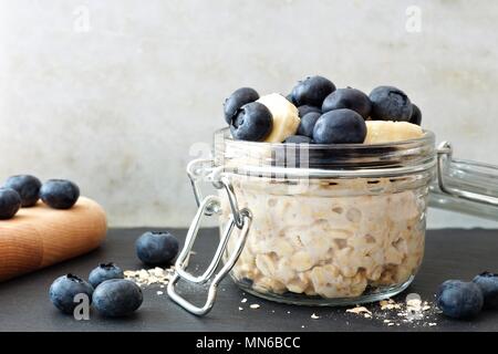 Glas über Nacht Hafer mit frischen Blaubeeren und Bananen, Szene mit weißen und schwarzen Stein Hintergrund Stockfoto