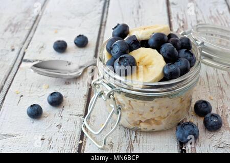 Über Nacht Hafer mit frischen Blaubeeren und Bananen in einem Glas auf einem rustikalen weiß Holz Hintergrund Stockfoto