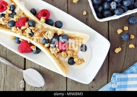 Gesunde Banana Split mit Joghurt, frischen Beeren und Müsli, über Szene auf Holz Stockfoto