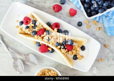 Gesunde Banana Split mit Joghurt, frische Beeren und Müsli, Overhead Szene auf Marmor Stockfoto