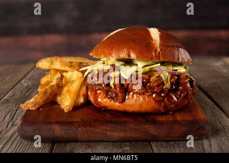 Zog pork Sandwich auf brezel Brötchen mit Kartoffelecken auf einer Platine mit Holz Hintergrund Stockfoto