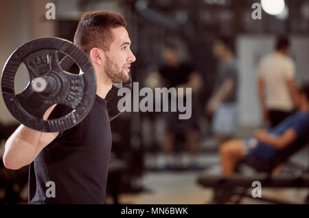 Obere Körper geschossen, Hocke übung Nahaufnahme, junger Mann mit Langhantel mit Gewichten auf dem Rücken Schultern. Nicht erkennbare Personen hinter der Turnhalle (unscharf) Stockfoto