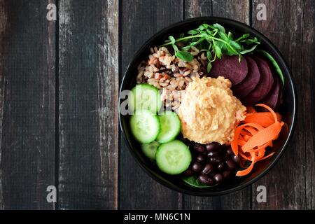 Gesunde vegetarische Ernährung Schüssel mit Hummus, Bohnen, wilder Reis, rote Rüben, Karotten, Gurken und Erbse Eintragfäden. Oben Blick auf dunklem Holz. Stockfoto