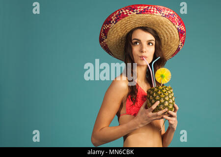 Junge schöne Frau in Bikini und Sombrero trinken Tropic cocktail Suchen auf blauem Hintergrund isoliert Stockfoto