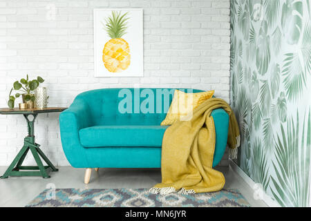 Gelbe Decke auf blauen Sofa neben dem grünen Tisch mit Werk in gemütliches Wohnzimmer mit Ananas Poster Stockfoto