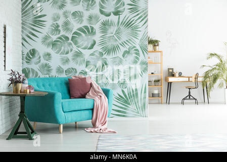 Rosa Decke und Kissen auf blauem Sofa neben einem Tisch mit Pflanzen im Zimmer mit Arbeitsbereich und geblümten Tapeten Stockfoto