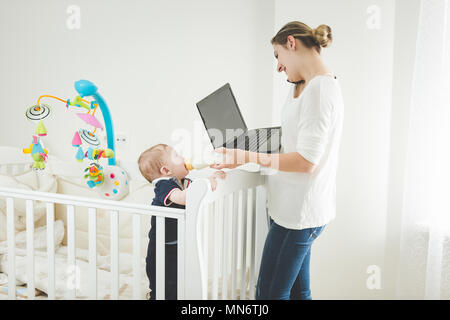 Junge Geschäftsfrau auf ocmputer zu Hause arbeiten und Ihr Baby Sohn Fütterung mit Milch Stockfoto