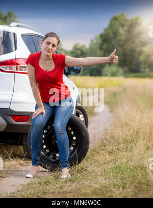 Verärgert junge Frau mit kaputten Auto halten Daumen hoch und Anhängevorrichtung - Wandern Stockfoto