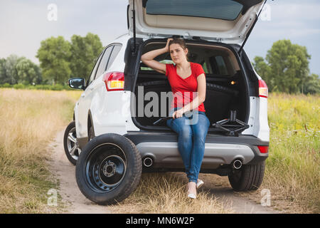Junge Frau sitzt im offenen Kofferraum und Haltewerkzeuge wor ändern Reifen Stockfoto
