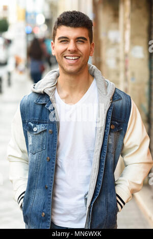 Gut aussehende junge Mann mit blauen Augen lächeln auf der Straße. Modell der Mode im städtischen Hintergrund tragen weiße T-Shirt, Jeans und blaue Jacke Stockfoto