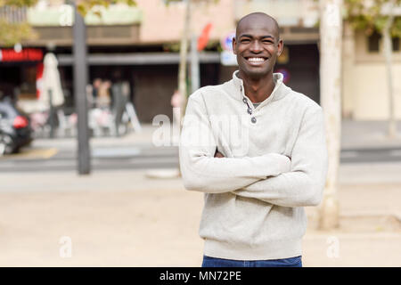 Schwarze jungen Mann mit verschränkten Armen lächelnd im städtischen Hintergrund. Junge afrikanische Kerl mit rasiertem Kopf legere Kleidung. Stockfoto