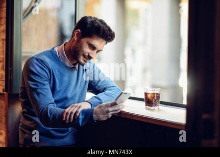 Junge lächelnde Mann mit blauen Pullover an seinem Smartphone in ein modernes Pub suchen. Kerl mit modernen Frisur eine Cola trinken Bärtigen. Stockfoto