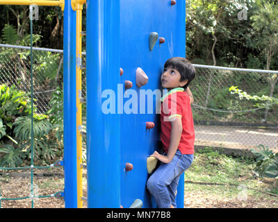 Kind bis auf die Kletterwand im Park. Junge Klettern bis auf Fahrt holding Griffe für Hände und Füße. Stockfoto