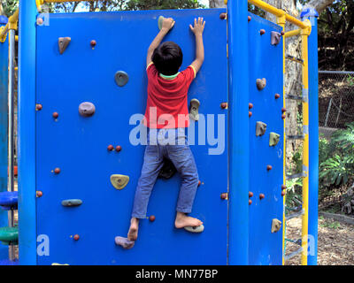 Kind bis auf die Kletterwand im Park. Junge Klettern bis auf Fahrt holding Griffe für Hände und Füße. Stockfoto