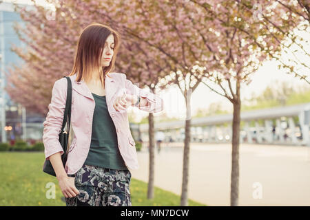 Junge Frau wartet auf die Besprechung oder Datum und Uhrzeit auf Armbanduhren. Schöne Frau unter blühenden sakura Bäume auf der Straße Stockfoto