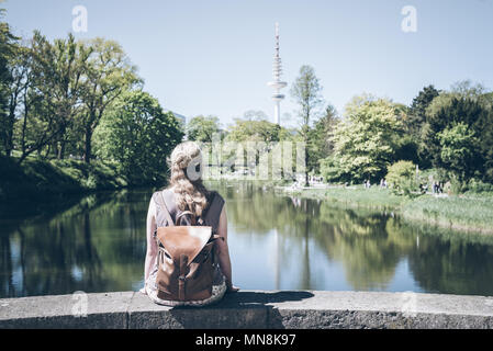 Rückansicht des blonden Frau sitzt auf der Wand mit Teich im Park an einem sonnigen Tag Stockfoto