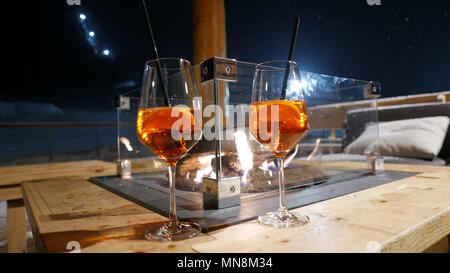 Zwei orange Weingläser mit Aperol Spritz Getränke und Strohhalme auf Winter Terrasse Tisch mit Kamin in der Mitte Stockfoto