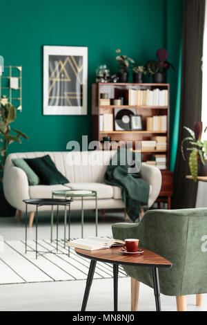 Holz- Tisch neben einem grünen Sessel im gemütlichen Wohnzimmer Interieur mit Poster an der Wand über dem Sofa Stockfoto