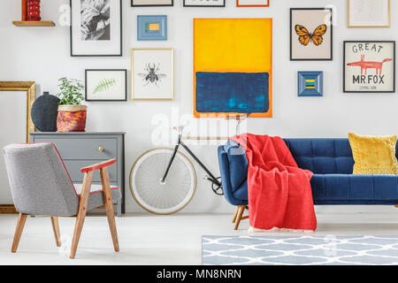 Rote Decke auf blauen Sofa im hellen Wohnzimmer Einrichtung mit gemusterten Sessel, Bike und viele Poster an die Wand geworfen Stockfoto